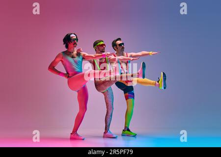 Drei Männer in klassischer, lustiger, farbenfroher Sportbekleidung, die sich vor einem abgestuften, blau-rosa Studiohintergrund streckt. Aerobic Stockfoto