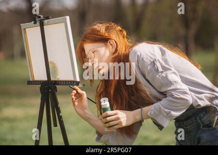 Nahaufnahme einer wunderschönen Künstlerin mit rotem Haargemälde auf einer Staffelei, während Sie im Park stehen Stockfoto