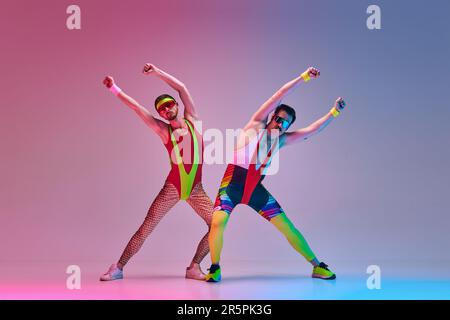 Junge Männer, Fitness-Coach in bunter Sportbekleidung, Stretching, Training vor einem abgestuften blauen, pinkfarbenen Studiohintergrund in Neonlicht Stockfoto