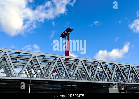 Nahaufnahme der Stahlbahnbrücke aus perspektivischer Sicht. Roter Stahlturm mit Sonnenkollektoren. Blauer Himmel mit weißen Wolken. Stabbalken mit dreieckigen Elementen Stockfoto