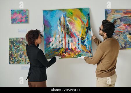 Ein paar Mitarbeiter hängen Gemälde an die Wand, sie bereiten sich auf die Kunstausstellung in der Galerie vor Stockfoto