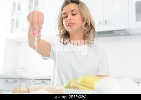 Ein junges Mädchen sitzt an einem Tisch in einer hellen weißen Küche und bereitet ein Mittagessen oder ein Gebäck-Dessert zu. Helle Farbtöne. Eine schöne weiße Blondine schüttet Mehl auf den Tisch. Vorderansicht mit Kopierbereich Stockfoto