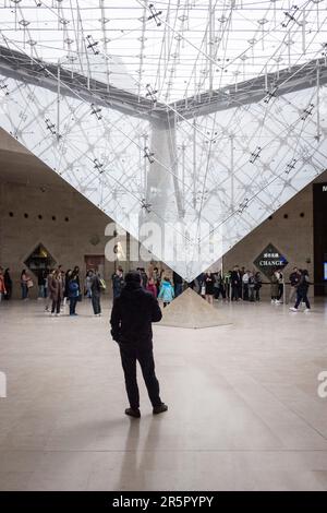 Die Menschen in Paris, Frankreich, versammeln sich um die fesselnde umgekehrte Pyramide, die sich im bekannten Einkaufszentrum „Carrousel du Louvre“ befindet, während sich die Menschen anstellen, um das Museum im Hintergrund zu betreten. Stockfoto