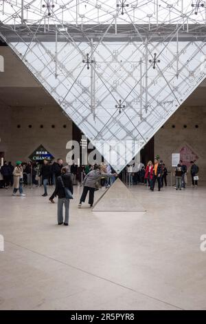 Die Menschen in Paris, Frankreich, versammeln sich um die fesselnde umgekehrte Pyramide, die sich im bekannten Einkaufszentrum „Carrousel du Louvre“ befindet, während sich die Menschen anstellen, um das Museum im Hintergrund zu betreten. Stockfoto