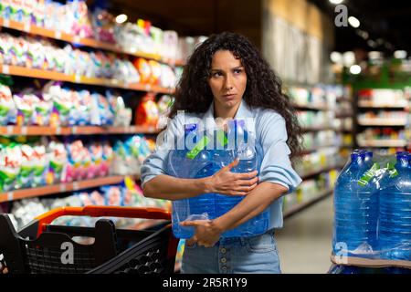 Eine junge Frau kauft Wasserpaletten in einem Supermarkt. Er nimmt es in die Hände, packt es in einen Wagen. Panik während einer Pandemie, Quarantäne Stockfoto