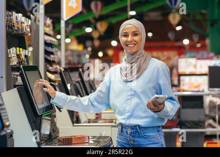 Selbstbedienung im Supermarkt. Eine junge arabische Frau in einem Hijab steht neben der elektronischen Kasse und bezahlt die Waren im Laden online über eine App auf ihrem Handy. Stockfoto