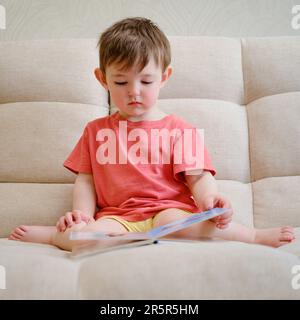 Das niedliche Baby spielt mit dem bunten Bilderbuch, blättert durch die Seiten und berührt die Bilder. Das kleine Kind ist in Gedanken verloren als Stockfoto