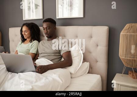 Ein junges, multiethnisches Paar, das verschiedene Geräte in seinem Bett benutzt Stockfoto