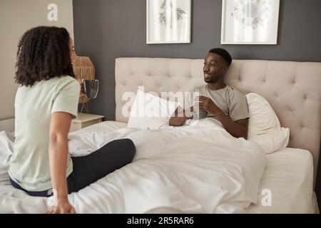 Lächelndes, junges, multirassisches Paar, das sich bei einem Kaffee im Bett unterhält Stockfoto