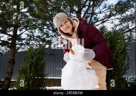 Eine schöne, fröhliche junge Frau mit Ohrenschützern macht einen Schneemann, der Spaß in einer schneebedeckten Natur hat. Winterspaß Stockfoto