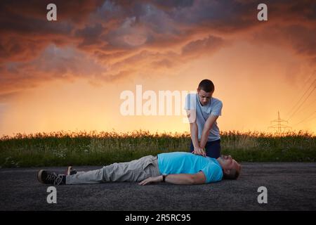 Dramatische Wiederbelebung auf der Landstraße. Ein junger Mann leistet erste Hilfe für einen bewusstlosen Mann mittleren Alters unter Sturmwolken. Die Themen Rettung, Hilfe und Hoffnung. Stockfoto