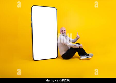 kaukasier mittleren Alters, der in der Nähe eines großen Smartphones für mobile Werbung sitzt. Neue Anwendung präsentieren, Mann zeigt Daumen hoch Konzeptidee. Stockfoto