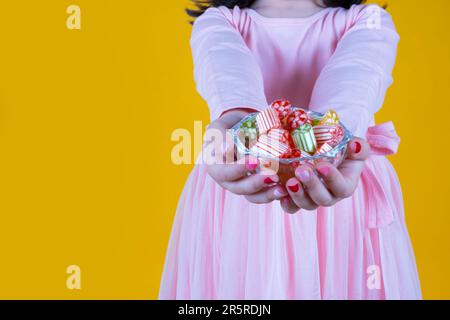 Kaukasisches kleines Mädchen, das harte Süßigkeiten hält und serviert. Schüssel mit buntem, köstlichem Süßes in einem Kristallglaskorb. Kind trägt pinkfarbenes Kleid. Stockfoto