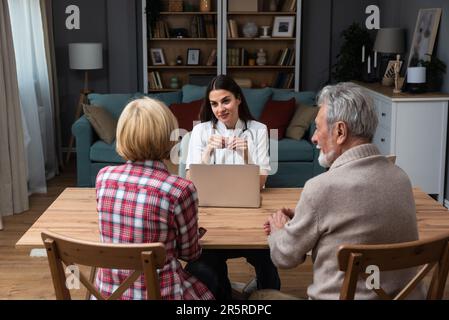 Weibliche Fachärztin, die Seniorenpaar-Patientin während des Arztbesuchs konsultiert. Junge Ärztin, die spricht und medizinische Hilfe leistet Stockfoto