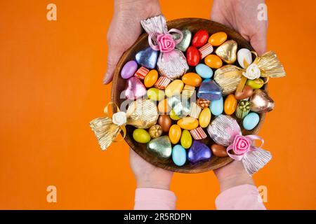 Die Hände von Frau und Kind halten eine Schüssel süß. Bild von oben: Umwickelte Luxus-Schokolade und Mandeldragee in Holzschale. Isolierter orangefarbener Hintergrund. Stockfoto