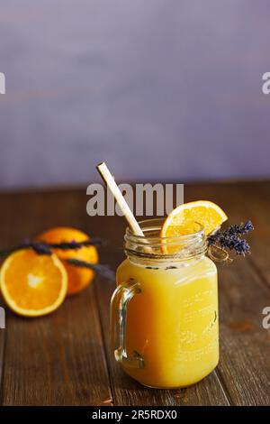 Orangefarbene Limonade in einem Becher auf einem Holztisch mit lavendelfarbenem Hintergrund Stockfoto