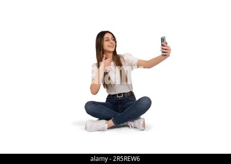Frau macht Selfie, Portrait der jungen kaukasierin, die Selfie macht. Auf dem Boden sitzend, mit Daumen-nach-oben-Geste. Mit dem Mobiltelefon. Stockfoto