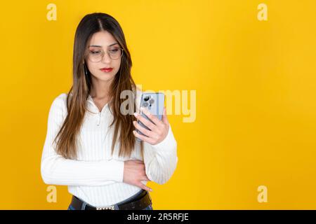 Eine ernste Frau. Geschäftsfrau steht auf isoliertem gelben Hintergrund, Kopierraum. Halten Sie Ihr Smartphone, sehen Sie den Bildschirm aus. Schlechte Nachrichten, unglücklich. Stockfoto