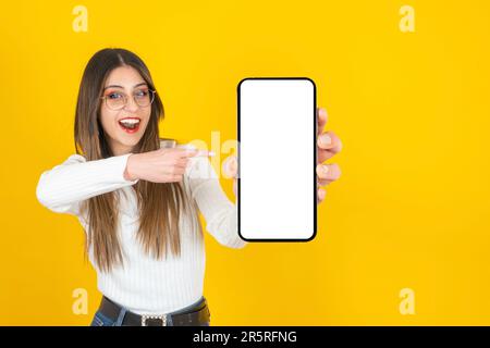 Ein weißes Mädchen, das mit dem Finger auf das Smartphone in der Hand zeigt. Leeres Mobiltelefon-Modell mit weißem Bildschirm. Reaktion auf isolierten gelben Hintergrund der neuen App Stockfoto