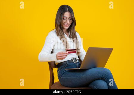 Frau sitzt auf einem Stuhl und sieht glücklich aus, mit Kreditkarte auf ihrer Hand zu stehen. E-Commerce, digitale Bezahlung, Bankgeschäfte, Ausgaben im Internet-Shop. Ich kaufe Waren. Stockfoto
