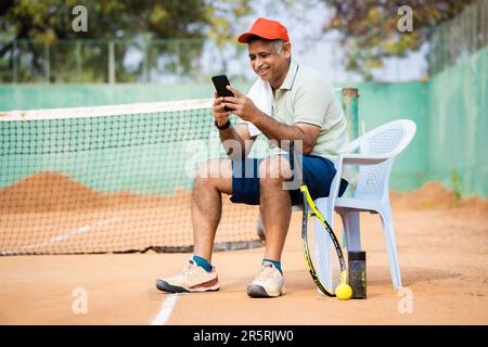 Glücklicher indain Senior Mann auf dem Tennisplatz, der während der Spielepause mit dem Handy beschäftigt ist - Konzept von Technik, sozialen Medien und Entspannung Stockfoto