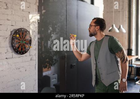 Fokussierter junger Mitarbeiter, der eine Pause bei der Arbeit hat und Darts spielt. Stockfoto