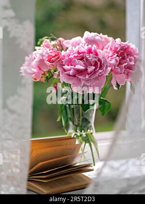 Ein Strauß schicke pinkfarbene Pfingstrosen in einer transparenten Glasvase und ein offenes Buch auf der Fensterbank Stockfoto