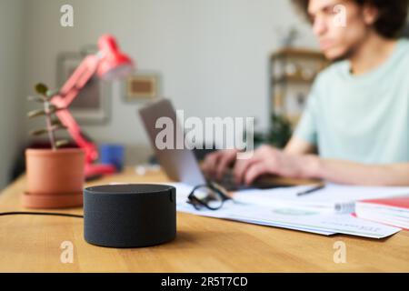 Nahaufnahme eines intelligenten Lautsprechers, der auf einem Tisch steht, während ein Mann im Hintergrund am Laptop arbeitet Stockfoto