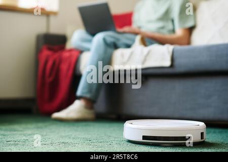 Nahaufnahme eines Roboter-Staubsaugers, der den Teppich saugt, während ein Mann am Laptop arbeitet, der im Hintergrund auf dem Sofa sitzt Stockfoto