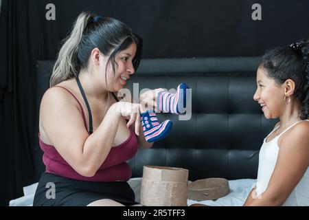 Junge Latina-Mutter zeigt ihrer kleinen Tochter Babyschuhe als Zeichen für eine Schwangerschaft. Stockfoto