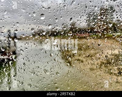 Eine Nahaufnahme von Regentropfen auf einem Glasfenster, mit einem Hintergrund mit üppigem grünen und braunen Gras und Bäumen Stockfoto