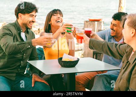 Eine vielfältige Gruppe von Freunden, die in einer Strandbar sitzen und fröhlich mit lebendigen Cocktails in Plastikbechern anstoßen. Die lebendige Szene zeigt die Freude Stockfoto