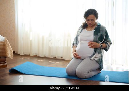 Wunderschöne Erwachsene schwangere Frau in Pose, hält ihren Bauch, macht Entspannungs- und Atemübungen auf der Fitnessmatte Stockfoto