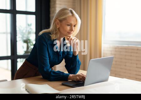 Attraktive, reife Geschäftsfrau, die zu Hause online an einem Laptop arbeitet, auf den Bildschirm schaut und denkt, freier Platz Stockfoto