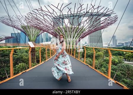 Eine lächelnde junge Frau in aufgewühltem Kleid auf einer erhöhten Plattform in Singapurs Gärten an der Bucht. Stockfoto