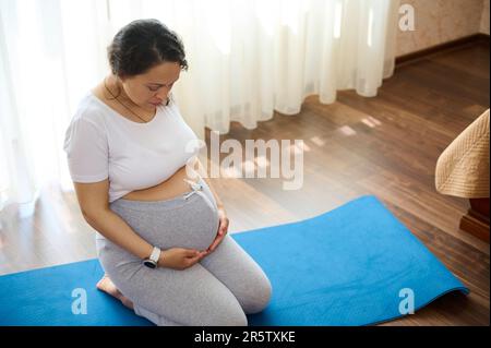 Draufsicht schwangere Frau hält ihre Hände am Bauch und übt Entspannungsübungen in heimeliger, ruhiger Atmosphäre Stockfoto