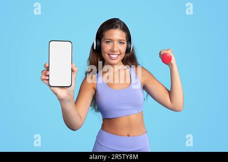 Fröhliche Millennial-Kaukasierin in Sportbekleidung in Kopfhörern mit Kurzhantel-Show-Telefon Stockfoto