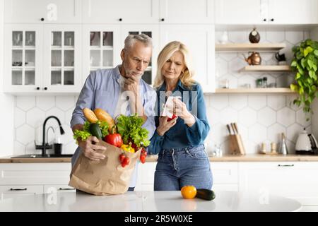 Essenskosten. Gestresstes Seniorenpaar In Der Küche, Das Nach Dem Einkaufen Rechnungen Überprüft Stockfoto