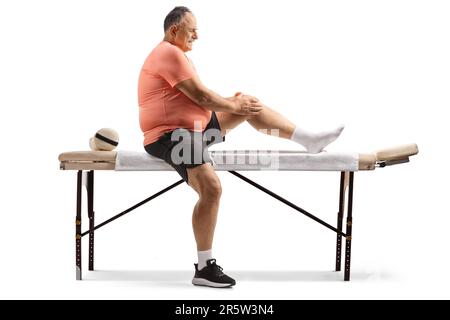 Profilaufnahme über die gesamte Länge eines Mannes, der auf einem Massagebett sitzt und sein Knie isoliert auf weißem Hintergrund hält Stockfoto