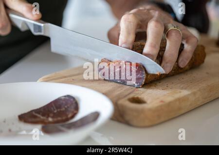 Ein Koch bereitet eine Mahlzeit in einer Küche zu und schneidet Pastirmastücke auf einem Holzbrett Stockfoto