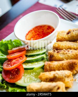 Vietnamesische Frühlingsrollen mit Tomaten, Gurken und roter Chilisauce auf dem Restauranttisch. Stockfoto