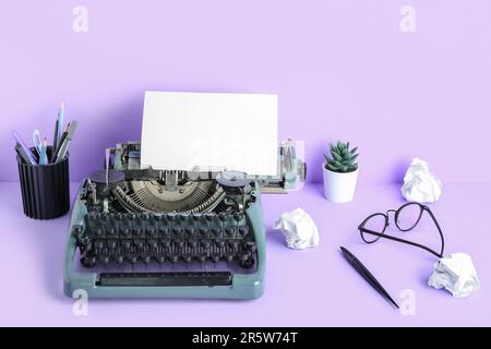 Vintage-Schreibmaschine mit Stifthalter, Brille und Hauspflanze auf lilafarbenem Hintergrund Stockfoto