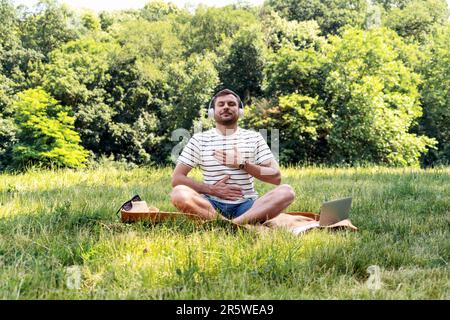 Ein Yoga-Mann, der im Sommer auf dem Rasen im Stadtpark sitzt und Meditationsmusik hört. Stockfoto
