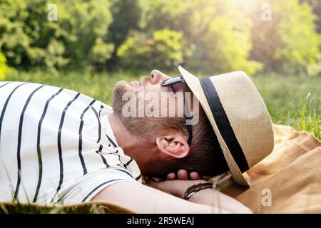 Ein Mann mit Sonnenbrille und Strohhut liegt auf dem grünen Rasen im Park und entspannt sich. Stockfoto