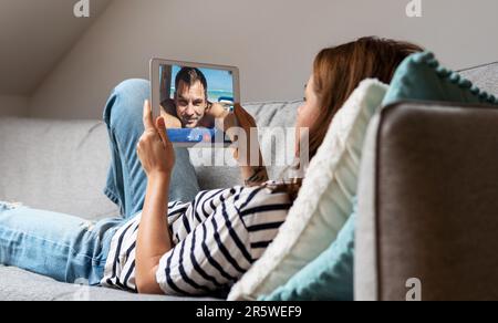 Junge Frau, die zu Hause auf dem Sofa liegt und ihren Freund per Video mit einem digitalen Tablet anruft. Stockfoto