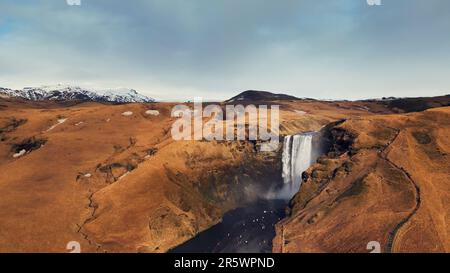 Drohnenaufnahme des nordischen Skgafoss Wasserfalls, der von gefrorenen Klippen herunterfällt und eine wunderschöne isländische Landschaft schafft. Riesige skandinavische Kaskade, die von farbenfrohen Hügeln abfloss. Zeitlupe. Stockfoto