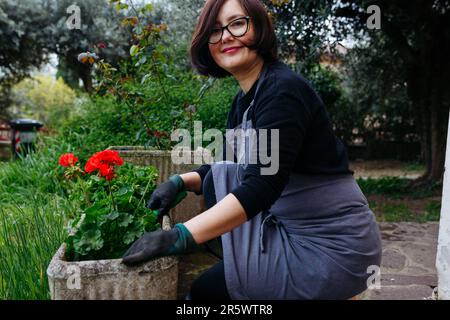Seitenansicht einer dunkelhaarigen Frau, die im Garten eine Geraniumblume in einen Topf pflanzt Stockfoto