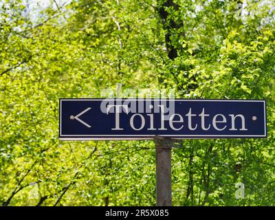 Ein Straßenschild mit Toiletten auf der linken Seite mit grünen, üppigen Bäumen im Hintergrund Stockfoto