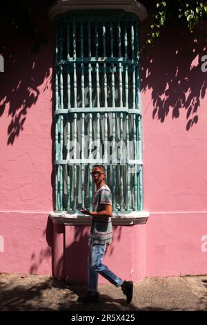 Cartagena, Provinz Cartagena, Bolivar, Kolumbien - Februar 16 2023: Junger Mann geht vorbei an einem rosafarbenen Haus mit einem riesigen Aqua Green Window, umgeben von Bou Stockfoto