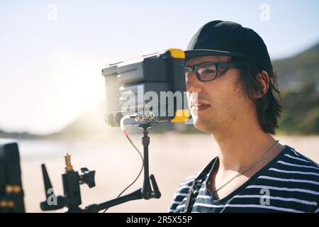 Seine Augen sind fest am Tatort. Ein fokussierter junger Mann, der tagsüber eine Szene mit einer hochmodernen Videokamera draußen am Strand dreht. Stockfoto